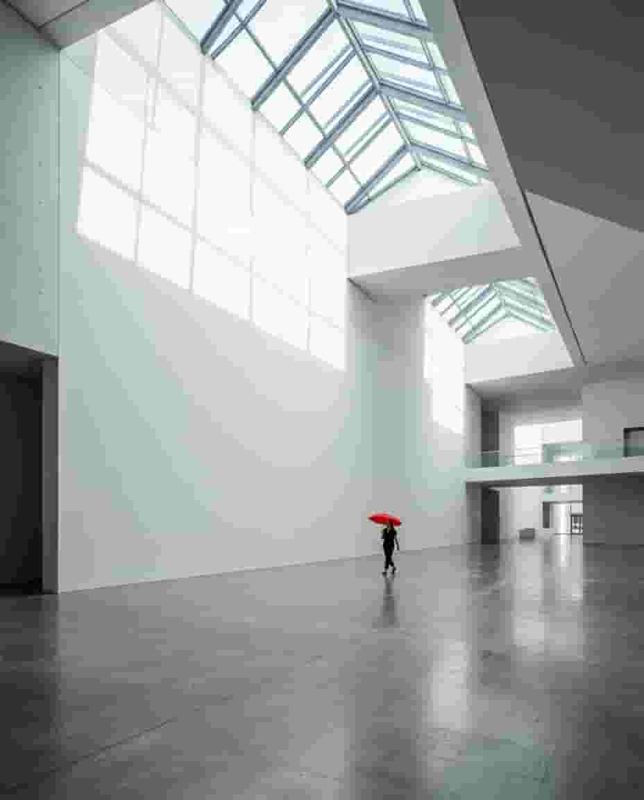 Bembeyaz minimalist bir modern sanat müzesinin devasa boş duvarı önünde kıpkırmızı şemsiyeyle yürüyen tek kişi, tepeden doğal ışık, çarpıcı negatif alan