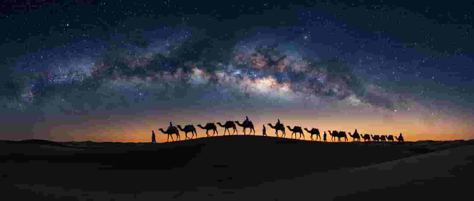 Muhteşem Samanyolu altında çöl kumullarını aşan deve kervanı silüetleri, yıldızlarla dolu lacivert gece, ufukta hafif sıcak ışıma, uzun pozlama astrofotoğraf